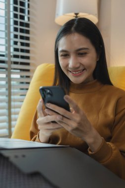 Güzel bir Asyalı kadın boş vaktini oturma odasında geçiriyor, akıllı telefonuyla oynuyor, hafta sonları evinde en sevdiği aktiviteleri yapıyor, rahat hissediyor ve eğleniyor.