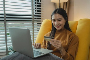 Oturma odasındaki kanepede oturan Asyalı kadın, kredi kartıyla alışveriş yapıyor, online alışveriş yapıyor, kredi kartı veya banka kartıyla ödeme yapıyor..