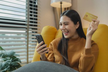 Oturma odasındaki kanepede oturan Asyalı kadın, kredi kartıyla alışveriş yapıyor, online alışveriş yapıyor, kredi kartı veya banka kartıyla ödeme yapıyor..