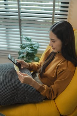 Oturma odasındaki kanepede oturan Asyalı kadın, kredi kartıyla alışveriş yapıyor, online alışveriş yapıyor, kredi kartı veya banka kartıyla ödeme yapıyor..