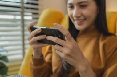 Güzel bir Asyalı kadın boş vaktini oturma odasında geçiriyor, akıllı telefonuyla oynuyor, hafta sonları evinde en sevdiği aktiviteleri yapıyor, rahat hissediyor ve eğleniyor.