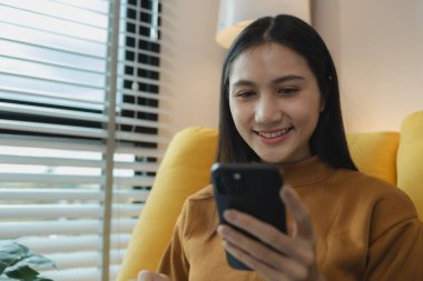 Güzel bir Asyalı kadın boş vaktini oturma odasında geçiriyor, akıllı telefonuyla oynuyor, hafta sonları evinde en sevdiği aktiviteleri yapıyor, rahat hissediyor ve eğleniyor.