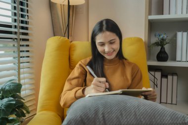 Asyalı bir kadın oturma odasındaki kanepede oturuyor. Geçmiş anılarını ve deneyimlerini kaydetmek için günlük tutuyor. Günlük tutmak boş zamanlarımızda bazı şeyleri düşünmemizi sağlayan bir aktivitedir..