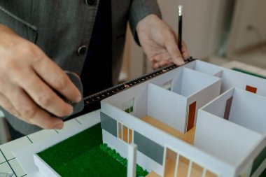 Ofiste çalışan bir mimar, bir mühendis bir ev modelinin ölçümlerini almak için bir araç kullanıyor. Böylece tasarıya uyuyor. Mimarın masasında bir ev modeli var.