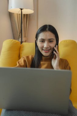 Güzel bir Asyalı kadın yurtdışındaki arkadaşlarıyla konuşmak için bir iletişim cihazı kullanıyor, evdeki kanepede dinleniyor ve kendi eğlence aktivitelerini yapıyor..