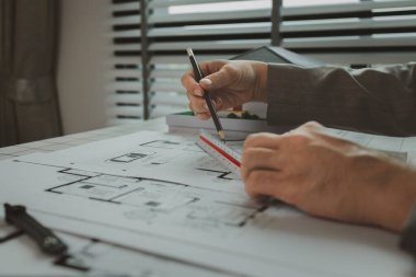 Ofiste bir mimar çalışıyor. Masanın üzerinde planlar ve ev modelleri var. Mühendis son bir kez daha mevcut olacak işleri kolaylaştırmak için araç kullanıyor.