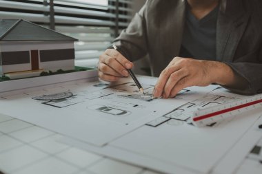 Ofiste bir mimar çalışıyor. Masanın üzerinde planlar ve ev modelleri var. Mühendis son bir kez daha mevcut olacak işleri kolaylaştırmak için araç kullanıyor.