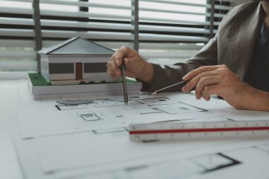 Ofiste bir mimar çalışıyor. Masanın üzerinde planlar ve ev modelleri var. Mühendis son bir kez daha mevcut olacak işleri kolaylaştırmak için araç kullanıyor.