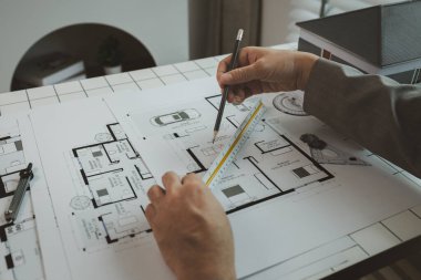 Ofiste bir mimar çalışıyor. Masanın üzerinde planlar ve ev modelleri var. Mühendis son bir kez daha mevcut olacak işleri kolaylaştırmak için araç kullanıyor.