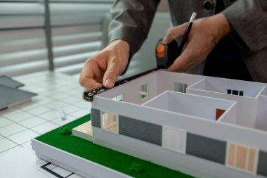 Ofiste çalışan bir mimar, bir mühendis bir ev modelinin ölçümlerini almak için bir araç kullanıyor. Böylece tasarıya uyuyor. Mimarın masasında bir ev modeli var.