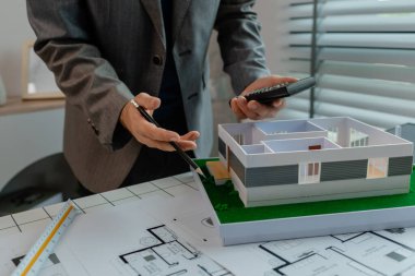 Bir mühendis, bir ev modelinin yapısal oranının hesap makinesi ile doğru olup olmadığını hesaplıyor, bir mimar, bir plandan rakamları kontrol etmek için hesap makinesi kullanıyor