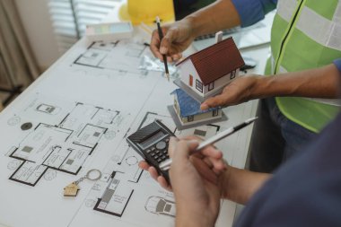 İki mimar ofiste ve birbirleriyle strateji planlarını paylaşıyorlar. Ofisteki masada iki mühendisle buluşup, planlarını ve ev modellerini paylaşıyorlar.