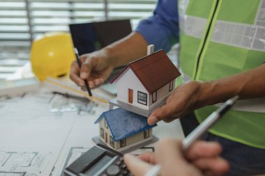 İki mimar ofiste ve birbirleriyle strateji planlarını paylaşıyorlar. Ofisteki masada iki mühendisle buluşup, planlarını ve ev modellerini paylaşıyorlar.