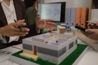 Ofiste bir mimar ekibiyle birlikte çalışıyor, planlar masanın üzerinde, müteahhit mühendislik ve mimari ile çalışıyor, ev modeli de masanın üstünde.