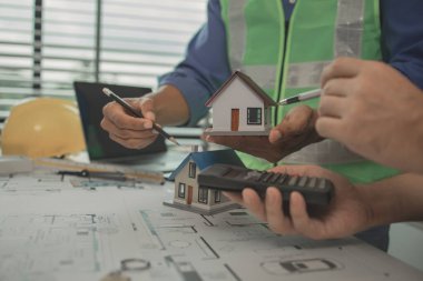 İki mimar ofiste ve birbirleriyle strateji planlarını paylaşıyorlar. Ofisteki masada iki mühendisle buluşup, planlarını ve ev modellerini paylaşıyorlar.