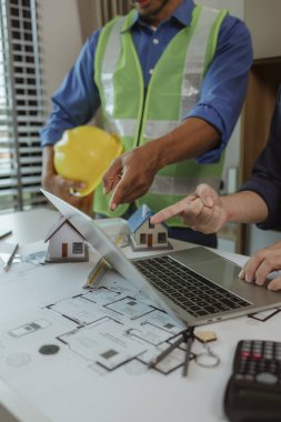 Bilimsel hesap makineleri, dizüstü bilgisayarlar, ev modelleri, ofisteki masanın üzerinde iki mimarla birlikte planlar var. Mühendis, müteahhitle talimat projesi hakkında görüşüyor.