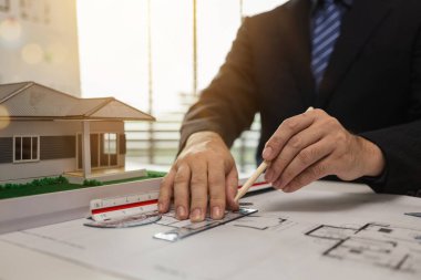 Ofiste çalışan bir mimar, işyerinde çalışan bir mühendis, plan ve ev modeli, çeşitli iş ekipmanlarıyla dolu bir mimarlık bürosu.