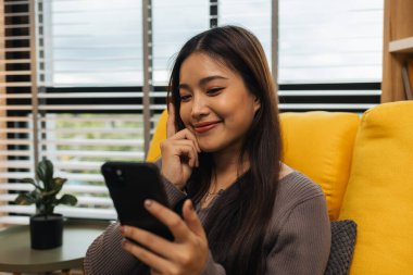 Gülümseyen genç bir kadın modern akıllı telefonu kullanıyor, çevrimiçi sohbet ediyor, evde dinlenen güzel bir kadın müzik dinliyor ve sevdiği diğer şeyler evde dinlenen düşünceli çok ırklı bir kadın.,