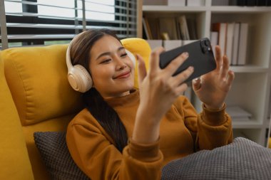 Gülümseyen genç bir kadın modern akıllı telefonu kullanıyor, çevrimiçi sohbet ediyor, evde dinlenen güzel bir kadın müzik dinliyor ve sevdiği diğer şeyler evde dinlenen düşünceli çok ırklı bir kadın.,