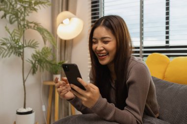 Gülümseyen genç bir kadın modern akıllı telefonu kullanıyor, çevrimiçi sohbet ediyor, evde dinlenen güzel bir kadın müzik dinliyor ve sevdiği diğer şeyler evde dinlenen düşünceli çok ırklı bir kadın.,