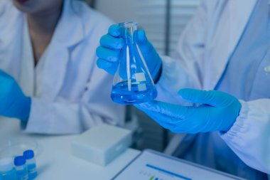 Laboratuvarda bilim adamları laboratuvar önlükleri, lastik eldivenler ve güvenlik gözlükleri takarken, iki kimyager sıvı haldeki kimyasalları test ediyor, kimyagerler laboratuarda çalışıyor.