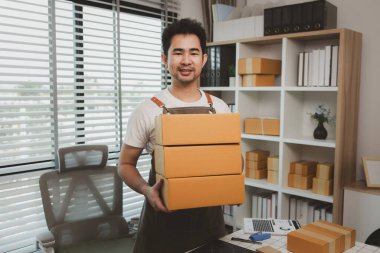 Elinde bir paket kutusu var ve kameraya bakıyor. Paket teslimatı için bir paket kutusu tutuyor. İnternetteki bir işyeri sahibi ofisinde.