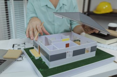 Asyalı erkek mimar çatı yapısını tutarken modern bir ev modelini analiz ediyor ve tasarımı bir kalemle işaret ediyor. Mimari planlama, mülkiyet tasarımı ve ev geliştirme kavramı.
