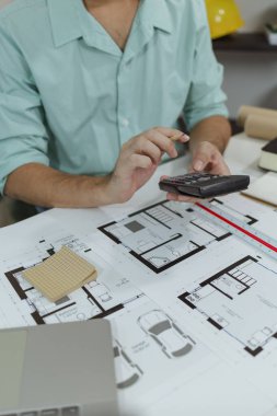 Yeşil gömlekli bir mimar beyaz bir masada oturuyor, basılı ev planlarını incelerken hesap makinesi kullanıyor. Resim çalışma alanı kurulumu ve mimari planlama sürecini vurgular.