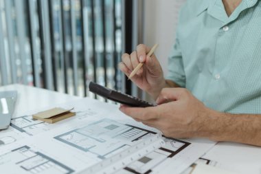 Yeşil gömlekli bir mimar beyaz bir masada oturuyor, basılı ev planlarını incelerken hesap makinesi kullanıyor. Resim çalışma alanı kurulumu ve mimari planlama sürecini vurgular.