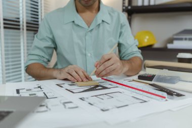 Masada mimari planlarla çalışan bir mimar, çizim ya da ayarlamalar için elinde bir kalem tutuyor. Tasarım planlama araçları ve ekipmanlarıyla profesyonel çalışma alanı.