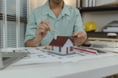 Bir mimar mimari planlara yerleştirilmiş bir ev modelini ölçmek için üçgen bir cetvel ve kalem kullanır, tasarım süreci sırasında oranları ve detayları kontrol eder.