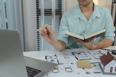 Ev tasarımı projesinde dizüstü bilgisayar kullanan, elinde kalem ve defter tutan bir mimar. Masada kat planları ve küçük bir ev modeli görülüyor. Detaylı planlama ve geliştirme çalışmaları öneriliyor.