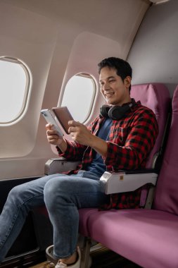 Asyalı adam pencerenin yanındaki uçak koltuğunda rahat bir şekilde oturuyor, elinde akıllı telefon ve pasaport, boynuna kulaklık takıyor, uçuş eğlencesinin tadını çıkarıyor. Modern seyahat yaşam tarzı ve teknoloji.