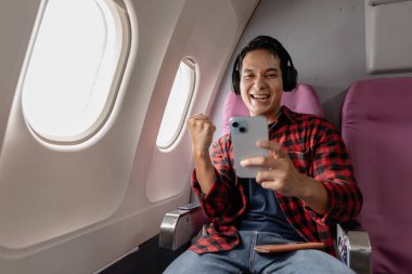 Uçakta neşeli bir genç adam akıllı telefonuna bakarken yumruğuyla kutlama yapıyor. Kulaklığı ve flanel gömleği ile yaklaşan yolculuğundan dolayı heyecanlı görünüyor..