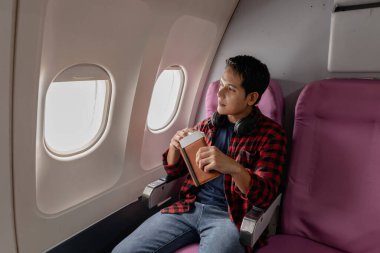 Düşünceli bir genç adam uçak koltuğunda oturmuş, elinde içinde para olan bir pasaport tutarken pencereden dışarı bakıyor. Sakin ve kendinden emin bir şekilde bir sonraki yolculuğuna hazır görünüyor..