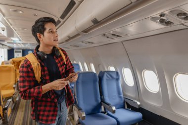 Asyalı bir adam uçağa biniyor ve elinde pasaport ve uçuş kartıyla koltuğunu arıyor. Sırt çantası taşırken ve boynunda kulaklık varken mutlu ve heyecanlı görünüyor..