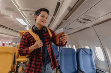 Asyalı bir adam uçağa biniyor ve elinde pasaport ve uçuş kartıyla koltuğunu arıyor. Sırt çantası taşırken ve boynunda kulaklık varken mutlu ve heyecanlı görünüyor..