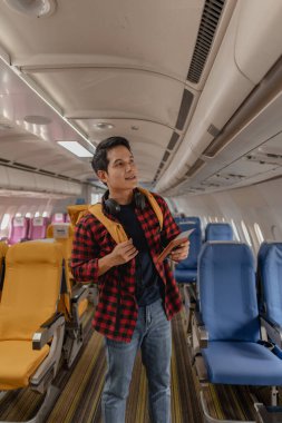 Asyalı bir adam uçağa biniyor ve elinde pasaport ve uçuş kartıyla koltuğunu arıyor. Sırt çantası taşırken ve boynunda kulaklık varken mutlu ve heyecanlı görünüyor..