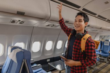 Asyalı bir adam uçağa biniyor ve elinde pasaport ve uçuş kartıyla koltuğunu arıyor. Sırt çantası taşırken ve boynunda kulaklık varken mutlu ve heyecanlı görünüyor..