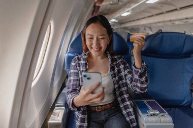 Uçağın penceresinde oturan, elinde kredi kartı ve akıllı telefonuyla gülümseyen mutlu genç bir kadın. Seyahati ve uçuşu sırasında eğlendiği için heyecanlı görünüyor..