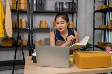 Genç bayan işyeri sahibi evde mutlu bir şekilde çalışıyor, çevrimiçi giyim mağazasını yönetiyor, siparişleri kontrol ediyor ve not alıyor, başarılı paketleme ve müşterilere teslimat için dizüstü bilgisayar kullanıyor..