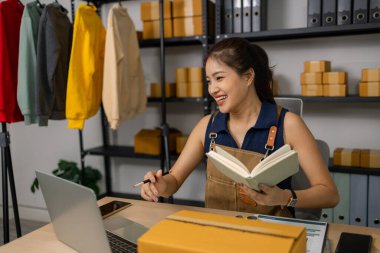 Genç bayan işyeri sahibi evde mutlu bir şekilde çalışıyor, çevrimiçi giyim mağazasını yönetiyor, siparişleri kontrol ediyor ve not alıyor, başarılı paketleme ve müşterilere teslimat için dizüstü bilgisayar kullanıyor..