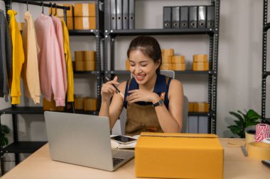 Genç bayan girişimci ev ofisinden online moda işini neşeyle yönetiyor, müşteri siparişlerini gözden geçiriyor ve dizüstü bilgisayar kullanarak teslimat için ürün paketleri hazırlıyor..