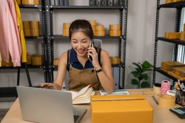 Gülümseyen genç bayan girişimci cep telefonuyla konuşuyor ve ev ofisinden dizüstü bilgisayarla çalışıyor, çevrimiçi giyim mağazasının müşteri siparişlerini yönetiyor ve sevkiyat ve teslimat için ürünler hazırlıyor..