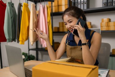 Gülümseyen genç bayan girişimci cep telefonuyla konuşuyor ve ev ofisinden dizüstü bilgisayarla çalışıyor, çevrimiçi giyim mağazasının müşteri siparişlerini yönetiyor ve sevkiyat ve teslimat için ürünler hazırlıyor..