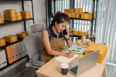 Gülümseyen genç bayan girişimci cep telefonuyla konuşuyor ve ev ofisinden dizüstü bilgisayarla çalışıyor, çevrimiçi giyim mağazasının müşteri siparişlerini yönetiyor ve sevkiyat ve teslimat için ürünler hazırlıyor..