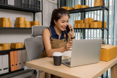 Gülümseyen genç bayan girişimci cep telefonuyla konuşuyor ve ev ofisinden dizüstü bilgisayarla çalışıyor, çevrimiçi giyim mağazasının müşteri siparişlerini yönetiyor ve sevkiyat ve teslimat için ürünler hazırlıyor..