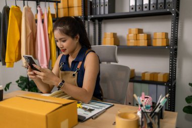 Genç bayan girişimci akıllı telefondan müşteri siparişlerini kontrol ediyor, çevrimiçi kıyafet işini mutlulukla yönetiyor, sevkiyatları organize ediyor ve ev ofisinde sipariş hazırlıkları için notlar alıyor..