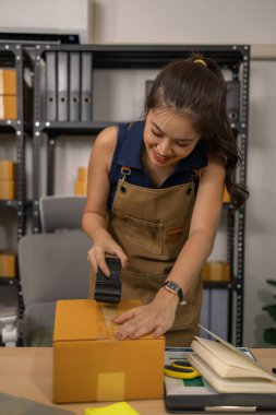 Genç bayan girişimci paketini bant ile dikkatlice mühürlüyor, mal siparişlerini hazırlıyor, başarılı online giyim işi için ev işindeki sevkiyat ve teslimatı yönetiyor..