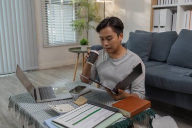 Genç bir adam evinde yerde oturmuş laptopuyla çalışırken belgeleri kontrol ediyor. Rahat ve modern bir yaşam alanında görevlerini yerine getirirken odaklanmış ve meşgul görünüyor..